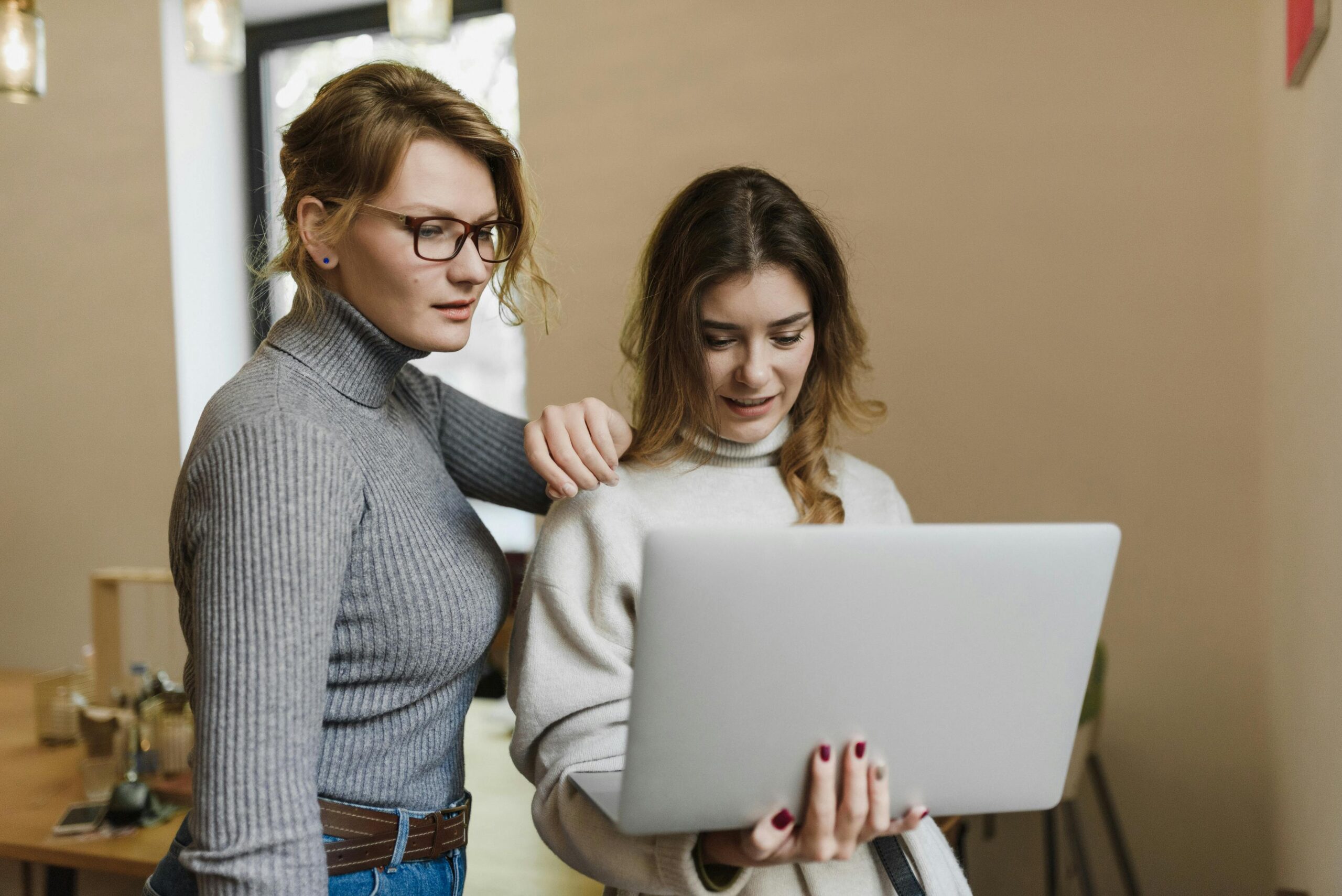 Two women in turtlenecks work together on a laptop in a warm indoor setting, showcasing teamwork.