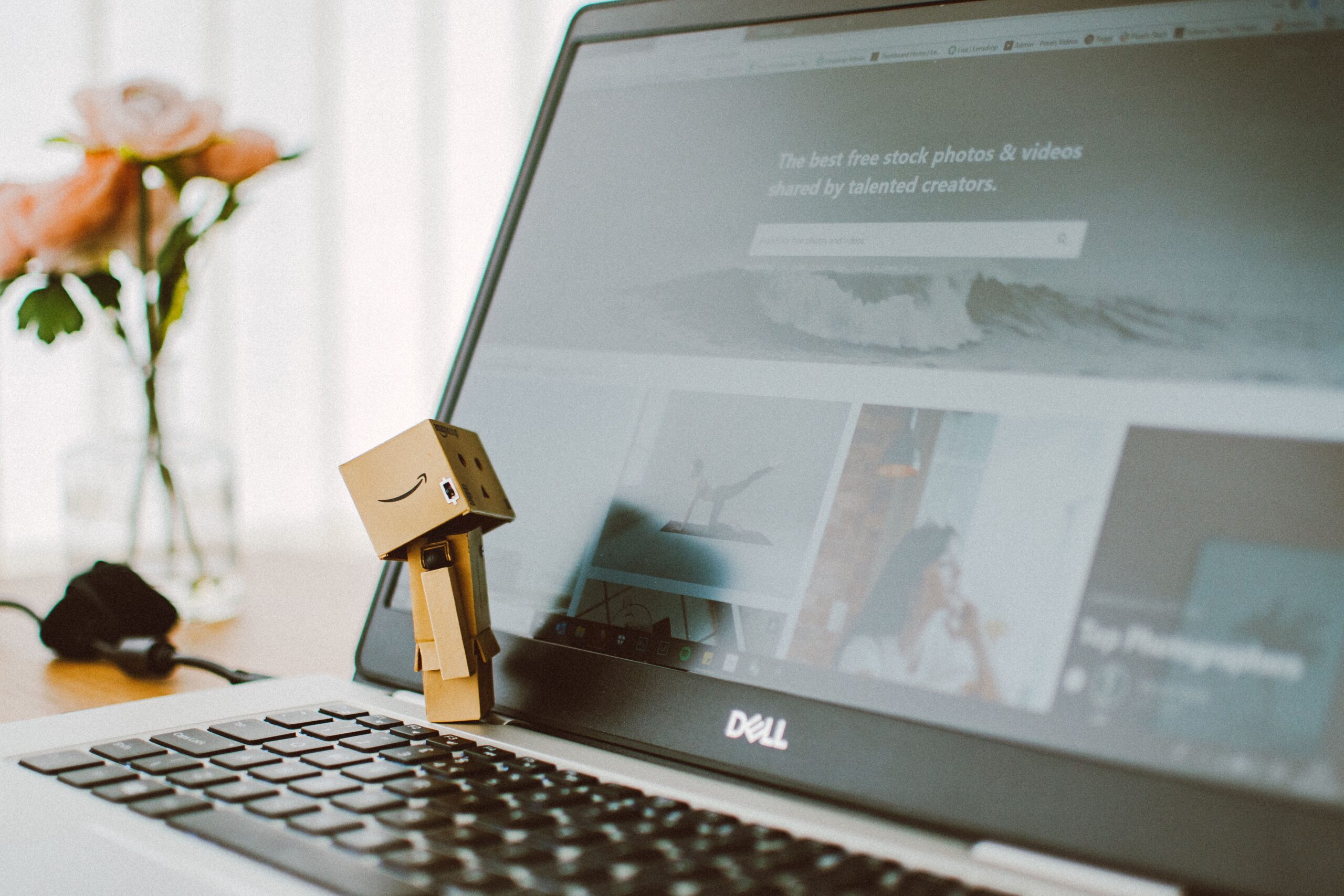 A small toy figure stands on a laptop keyboard with flowers in the background.